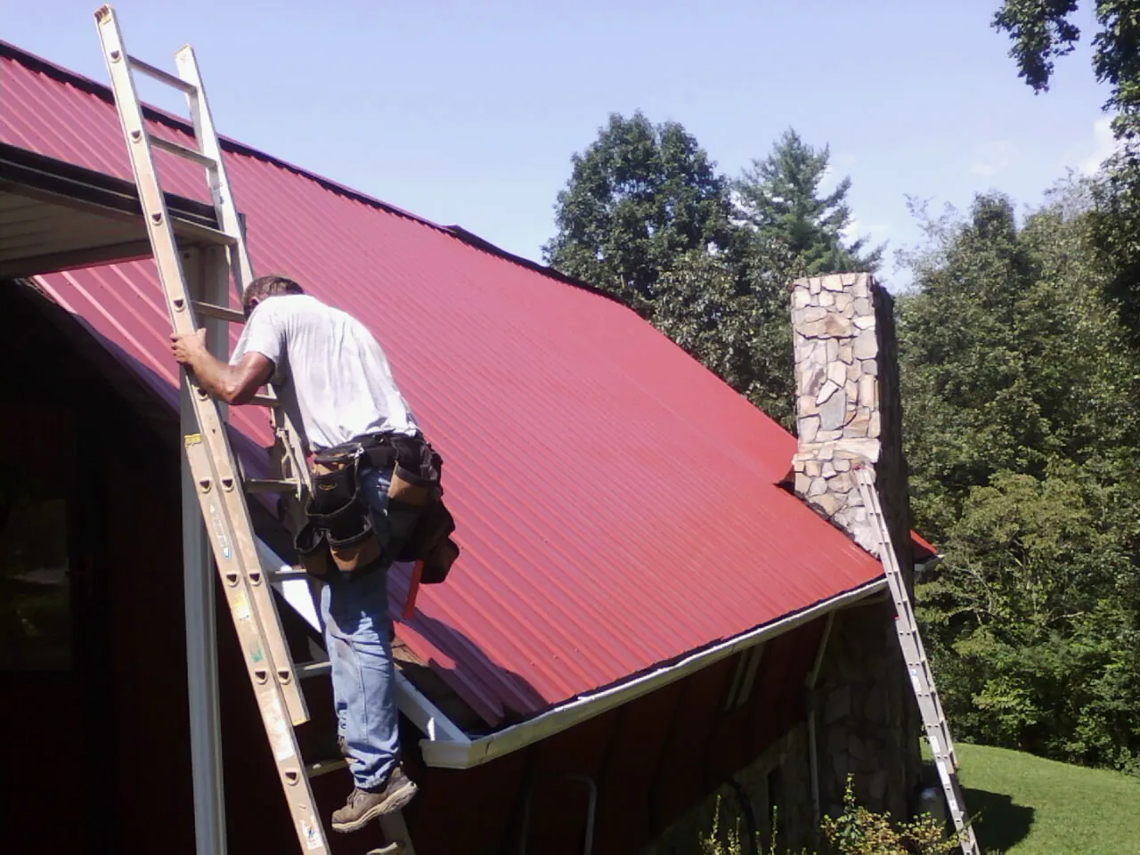 Roofer on a ladder beside a red metal roof and stone chimney — Soffit & Fascia Repair by Roofing Brothers in Blue Rock